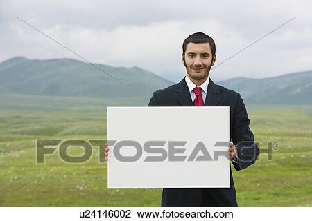 Smiling businessmen standing in mountain field holding blank sign front view View Large Photo Image Stock Image - Smiling businessmen standing in mountain field holding blank sign front view. Fotosearch