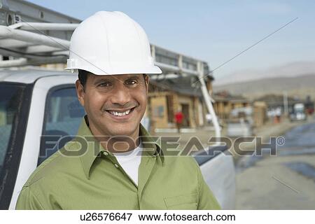 Stock Photo - Smiling Construction Worker in hard hat standing next to truck on construction Site. Fotosearch