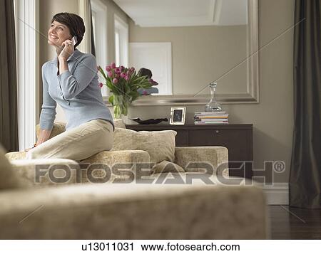 Stock Image - Smiling woman talking on mobile phone in living room. Fotosearch