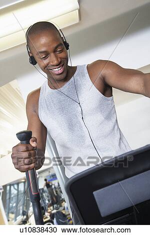 Stock Image - Sweaty man on elliptical machine in health club listening to headphones. Fotosearch