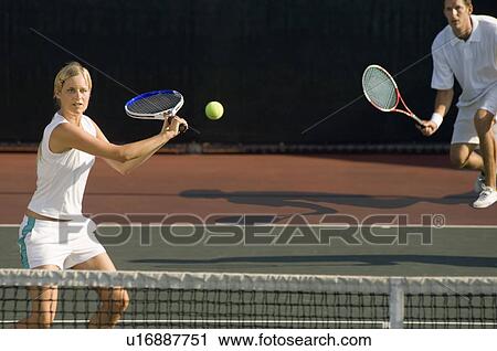 Stock Image - Tennis Player Swinging at Ball near tennis net; doubles partner squatting behind. Fotosearch