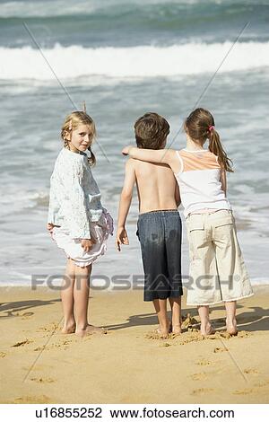 Three children standing at water edge on beach back view View Large Photo Image Stock Image - Three children standing at water edge on beach back view. Fotosearch