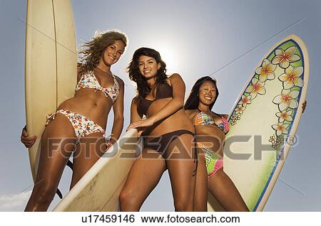 Stock Photograph - Three female surfers holding surfboards low angle view. Fotosearch