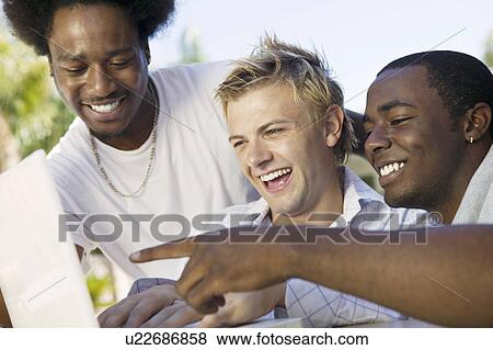 Stock Photo - Three friends at back yard table looking at laptop laughing close up. Fotosearch