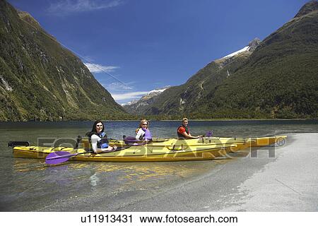 Stock Image - Three people in kayaks at shore of mountain lake. Fotosearch