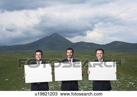 Stock Image - Three smiling businessmen standing in mountain field holding blank signs front view. Fotosearch