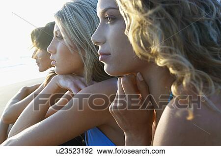 Three young women sitting side by side on beach looking at view close up View Large Photo Image Stock Image - Three young women sitting side by side on beach looking at view close up. Fotosearch