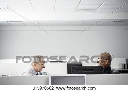 Stock Image - Two businessmen sitting face to face in office cubicle smiling side view. Fotosearch