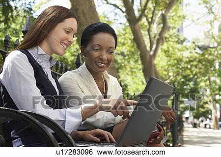 Two female colleagues using laptop on park bench View Large Photo Image Stock Photograph - Two female colleagues using laptop on park bench. Fotosearch