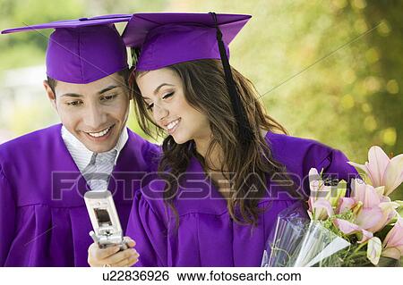 Two graduates looking at cell phone outdoors View Large Photo Image Stock Photograph - Two graduates looking at cell phone outdoors. Fotosearch