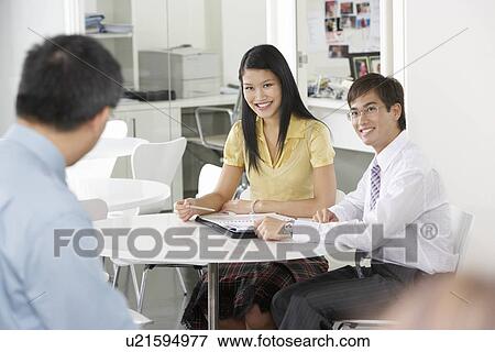 Two office workers sitting at table in office smiling at colleague View Large Photo Image Stock Photo - Two office workers sitting at table in office smiling at colleague. Fotosearch
