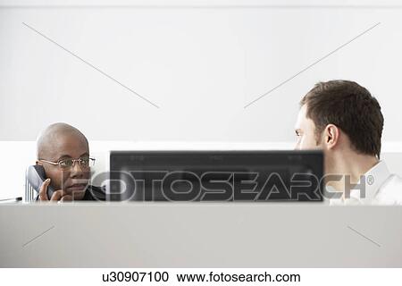 Stock Image - Two office workers sitting in office cubicle one using phone. Fotosearch