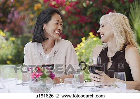 Stock Image - Two smiling women sitting at table holding wine glasses. Fotosearch