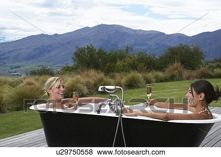 Stock Photo - Two women taking bath with champagne on porch near mountains. Fotosearch