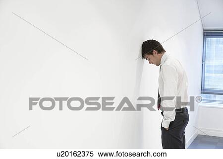 Stock Photography - Unhappy Businessman standing with head against wall in empty office. Fotosearch