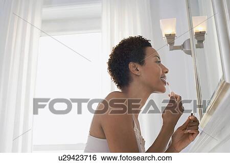 Stock Photograph - Woman applying lip gloss in mirror at home. Fotosearch