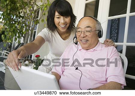 Stock Image - Woman assisting father wearing headset and using laptop on porch. Fotosearch