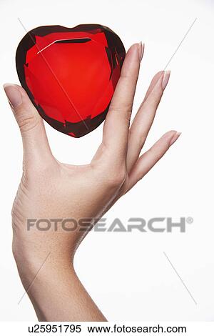 Stock Photography - Woman holding up heart-shaped jewel between finger and thumb close-up of hand. Fotosearch