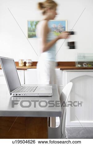 Stock Photograph - Woman in kitchen with coffee percolator walking by laptop focus on laptop. Fotosearch