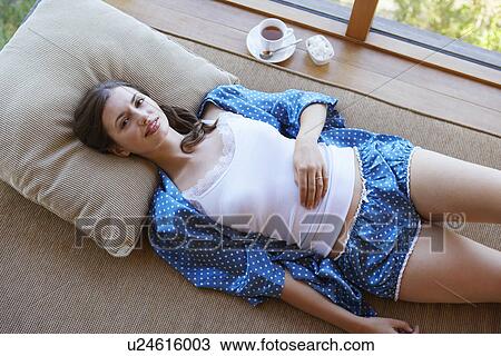 Stock Image - Woman in pajamas lying on couch with coffee on window sill. Fotosearch