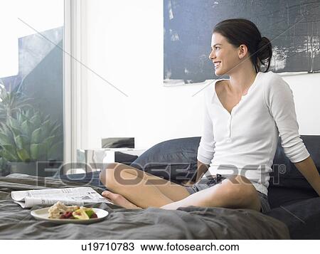 Stock Image - Woman sitting on bed with food and paper. Fotosearch