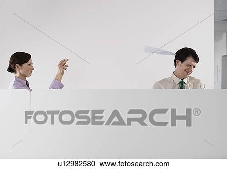 Stock Image - Woman throwing paper aeroplane at male colleague in office. Fotosearch