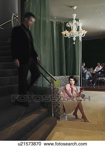 Stock Image - Woman waiting in hotel lobby for man descending staircase. Fotosearch