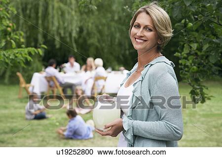 Stock Image - Woman with pitcher of lemonade in garden family members in background. Fotosearch