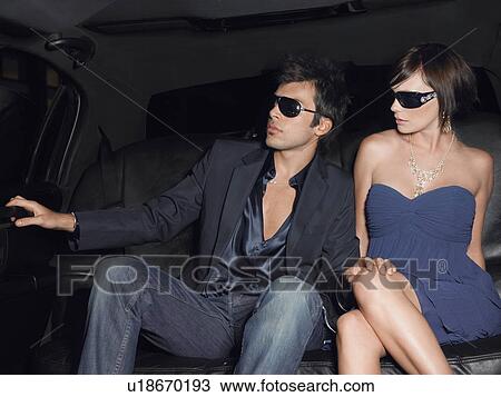 Stock Image - Young couple in backseat of limousine wearing evening dress and sunglasses. Fotosearch