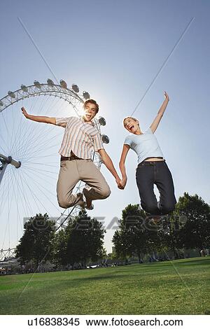 Stock Photography - Young couple in park jumping in air in front of London Eye portrait low angle view. Fotosearch
