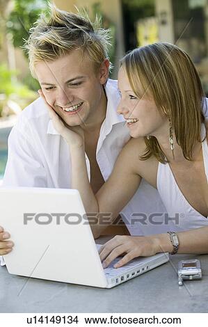 Picture - Young couple sitting at backyard table using laptop. Fotosearch
