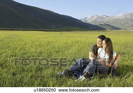 Stock Image - Young couple sitting in mountain field looking at view full length. Fotosearch