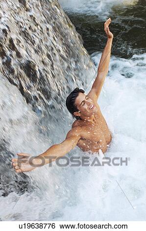 Stock Photograph - Young man standing under waterfall with arms outstretched high angle view. Fotosearch