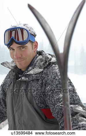 Picture - Young man wearing ski goggles on head holding skis in snow. Fotosearch
