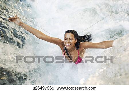 Stock Photography - Young woman standing under waterfall with arms outstretched high angle view. Fotosearch