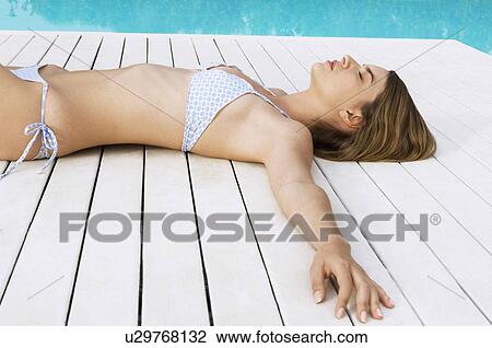 Stock Image - Young Woman Sunbathing on deck by swimming pool side view. Fotosearch