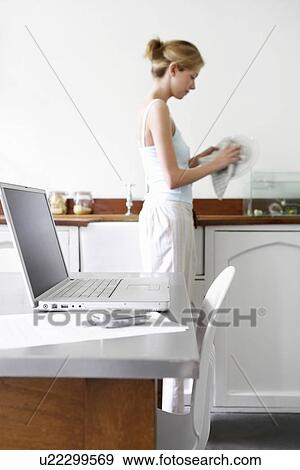 Stock Photo - Young woman washing dishes in kitchen by laptop and cell phone. Fotosearch