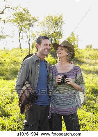 Picture - Adult couple outdoors man carrying blanket woman binoculars smiling. Fotosearch