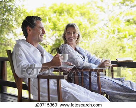 Stock Photo - Adult couple sitting on terrace man holding cup smiling. Fotosearch