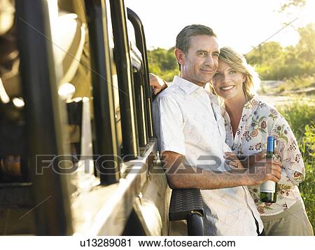 Stock Image - Adult couple standing by jeep man holding bottle of wine smiling. Fotosearch