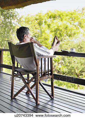 Picture - Adult man in bathrobe sitting on chair at terrace with feet up. Fotosearch