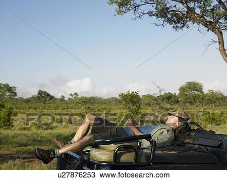 Stock Image - Adult man lying on bonnet of jeep having nap. Fotosearch