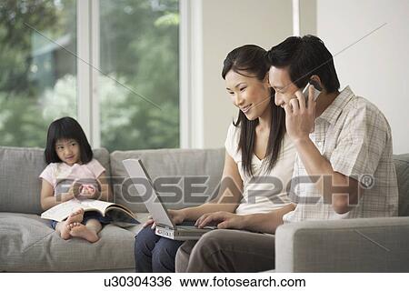 Stock Photograph - Asian couple using laptop on sofa with daughter watching in background. Fotosearch