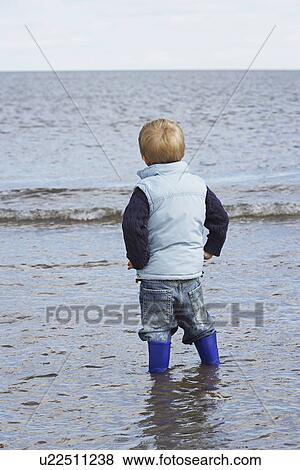 Stock Photo - Boy (3-4) wearing wellington boots standing in ocean. Fotosearch