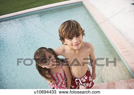 Stock Image - Boy (7-9) and girl (5-6) embracing at edge of swimming pool elevated view. Fotosearch