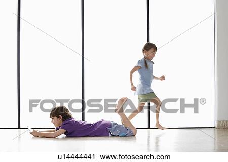Stock Image - Boy (7-9) lying on floor playing handheld game girl (5-6) running. Fotosearch