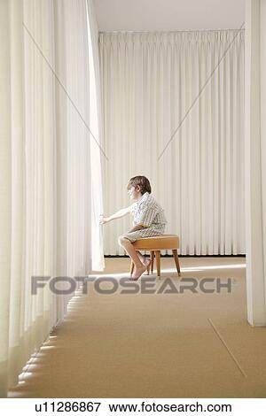 Boy (7-9) sitting on stool in room looking out window side view View Large Photo Image Stock Photo - Boy (7-9) sitting on stool in room looking out window side view. Fotosearch