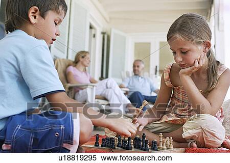 Stock Photography - Brother and sister (7-9) playing chess on porch parents in background. Fotosearch