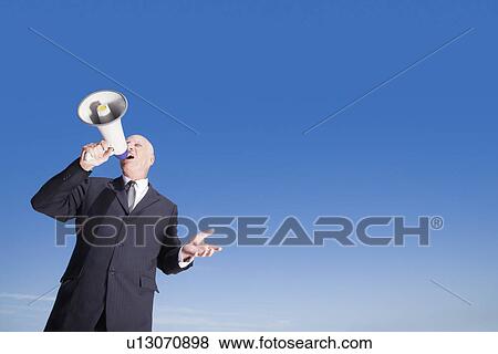 Stock Photo - Busieness man using megaphone standing against sky low angle view. Fotosearch