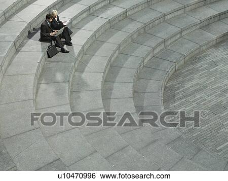 Stock Photograph - Business man and woman sitting on spiral stairs using laptop elevated view. Fotosearch